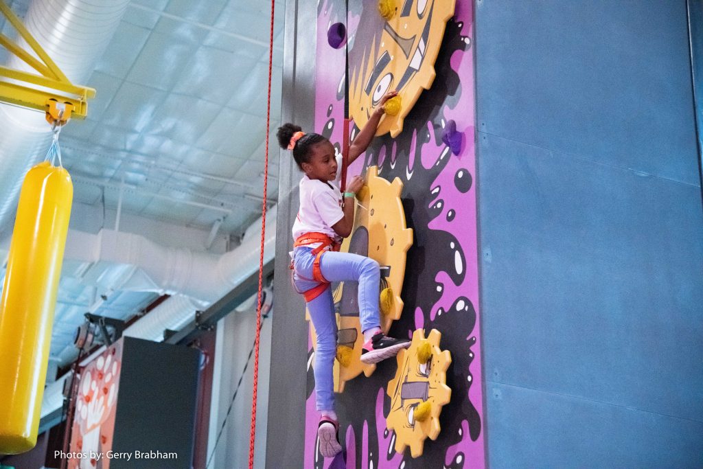 young girl in purple pants, white shirt, brown hair on a yellow and purple gear climbing wall